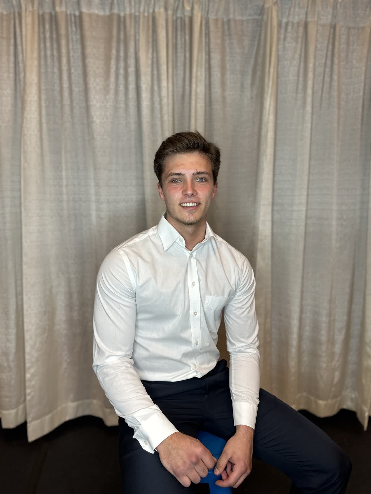 Matt Lyskawa seated and smiling, in a white dress shirt and dark trousers, in front of beige curtains.