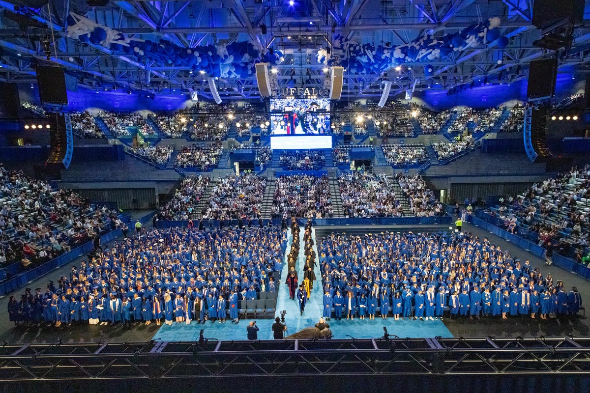Wide view of University at Buffalo commencement 2025, with graduates in blue caps and gowns filling an arena under blue lights.